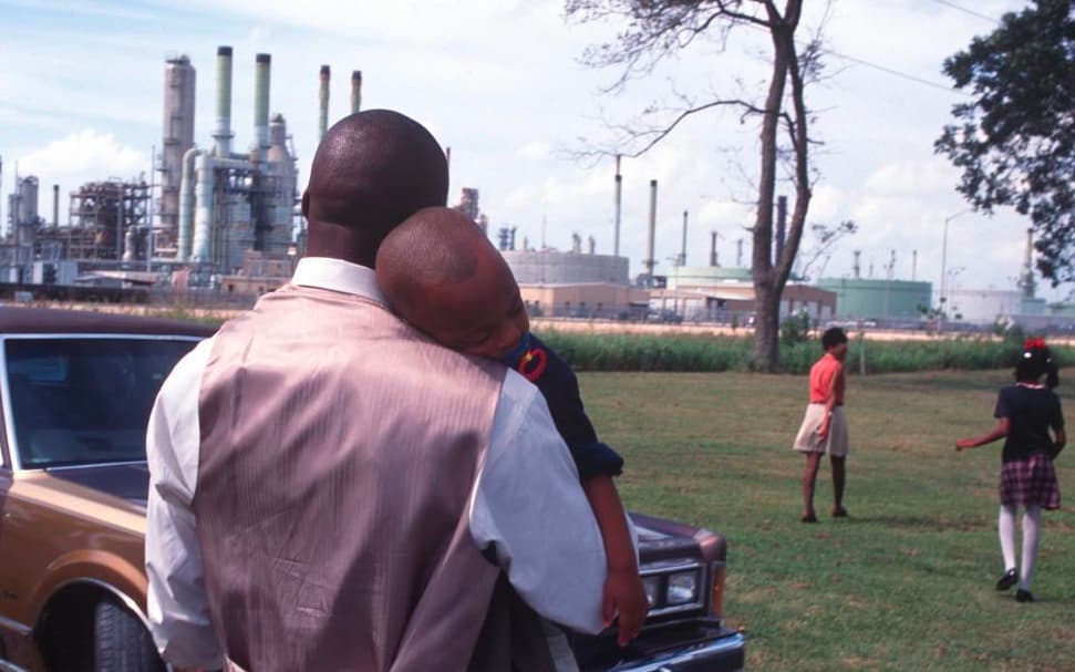 A father holds his child while two women stand nearby in a park beside an industrial facility